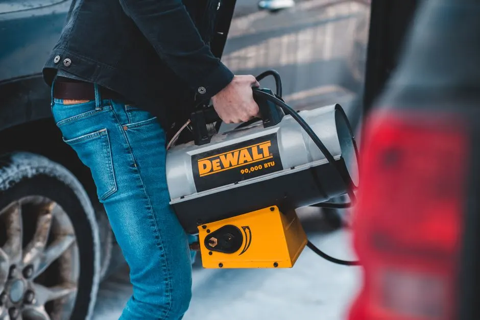 From above side view of crop unrecognizable slim male in jeans carrying portable blow heater with plastic handle and shiny metal tank standing near autos with frozen wheels